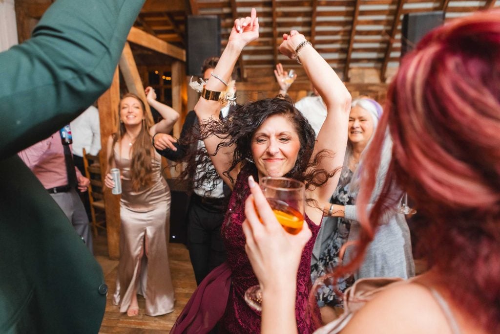 A group of people, including women and men, are dancing at a lively wedding reception. The woman in the center has her arms raised and is smiling, while another person with red hair holds a drink in the foreground. The Barns at Hamilton Station provides the perfect backdrop for this joyous celebration.