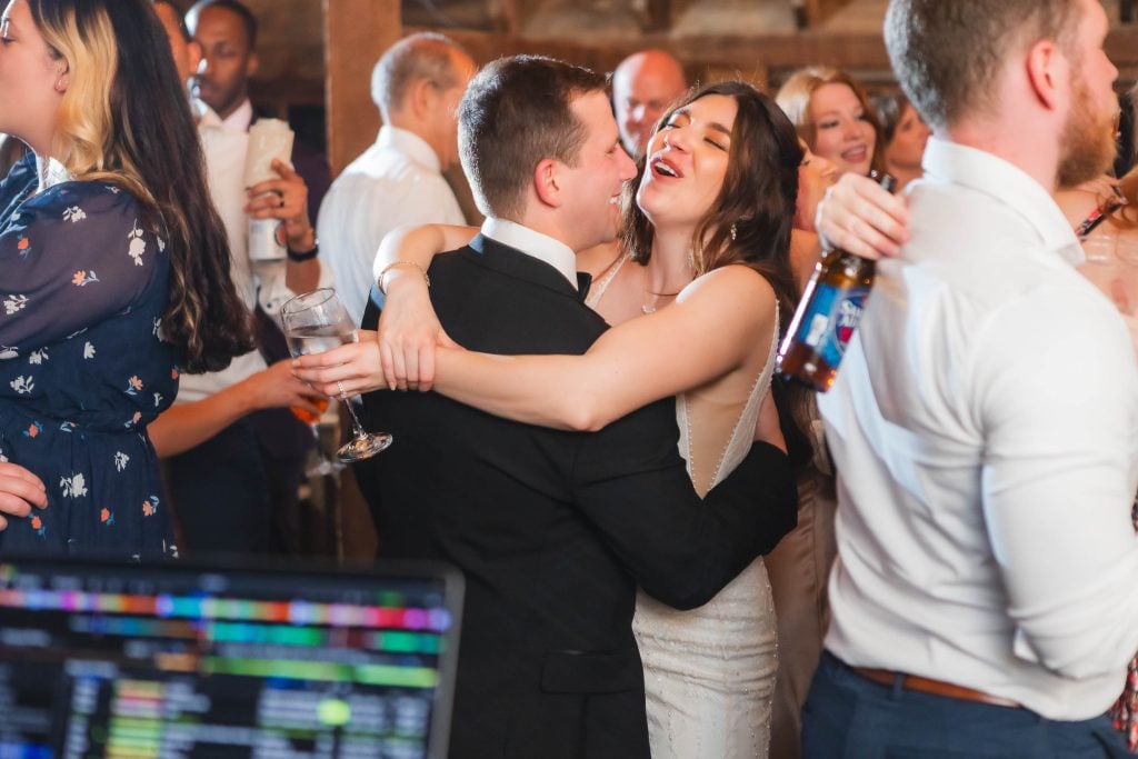 A couple embraces while dancing at a crowded indoor wedding reception at The Barns at Hamilton Station. The man holds a drink as they smile, and another man in the foreground holds a beer. A variety of people can be seen socializing in the background.