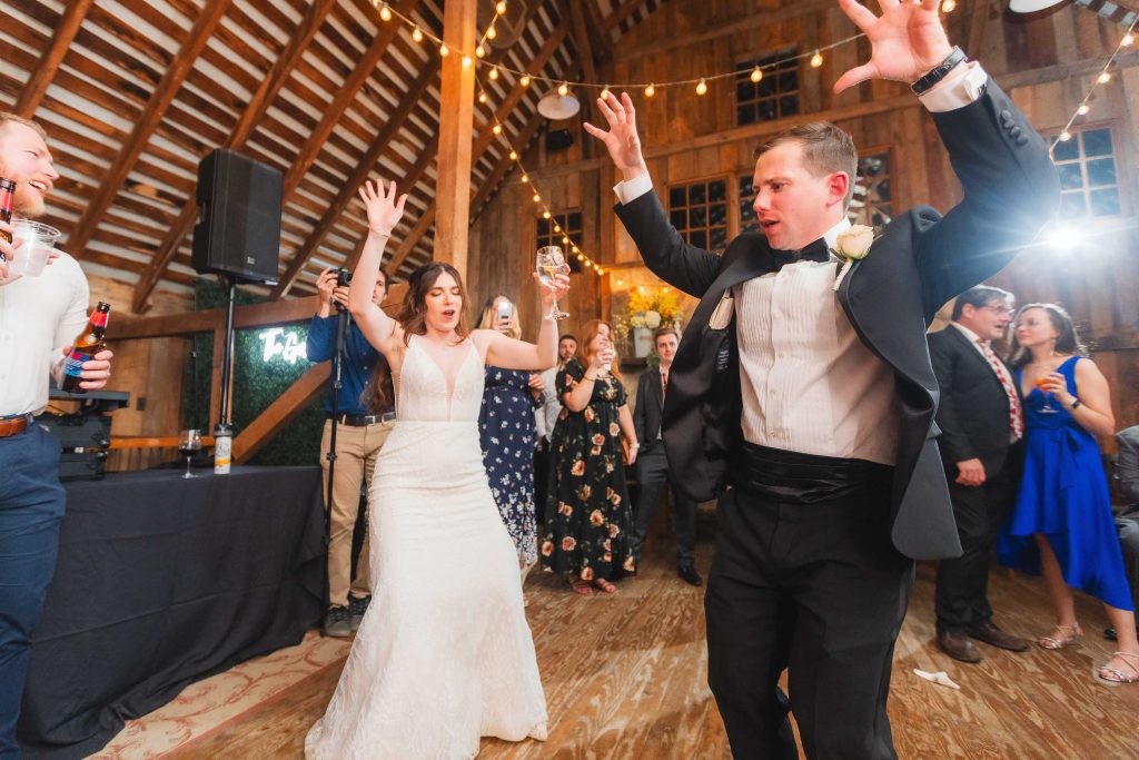 A bride and groom dance energetically at their wedding reception held at The Barns at Hamilton Station, while guests cheer and celebrate around them.