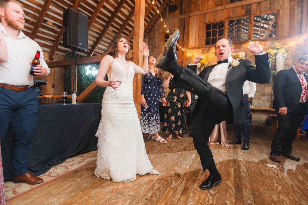 Bride and groom dancing energetically in The Barns at Hamilton Station, with guests standing nearby. The groom is kicking his leg up, and the bride is singing or speaking. Fairy lights decorate the wooden interior of the rustic venue, making this wedding reception unforgettable.