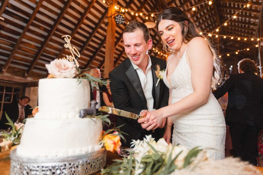 The bride and groom jointly cut a white tiered wedding cake in a warmly lit, decorated wooden venue at The Barns at Hamilton Station, setting the perfect tone for their beautiful wedding reception.