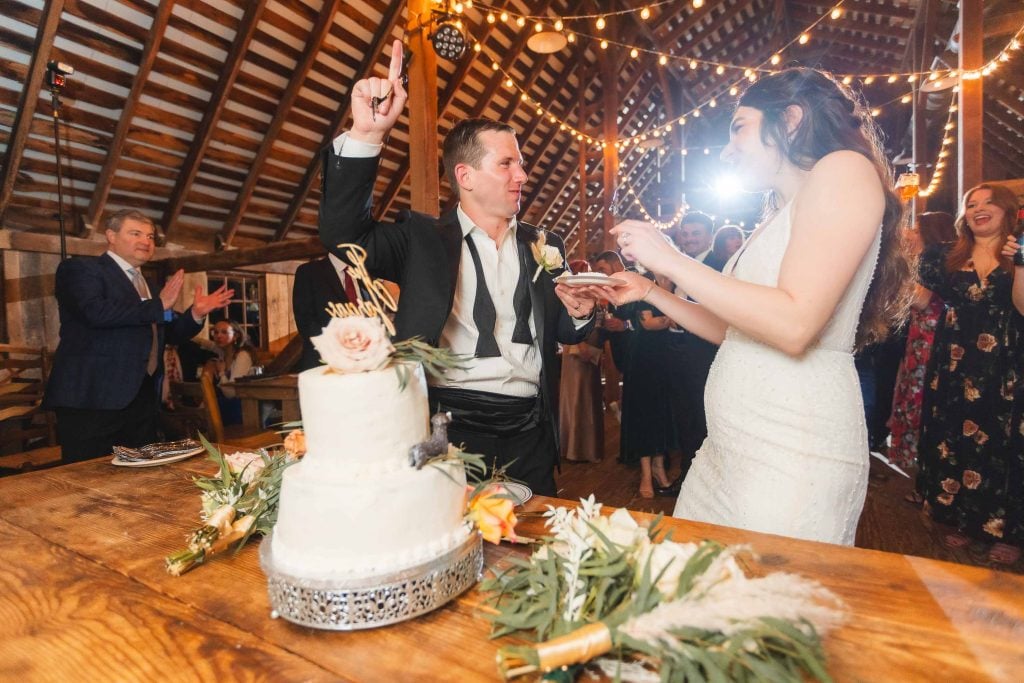 A newlywed couple is cutting a wedding cake at The Barns at Hamilton Station, a rustic, barnstyle venue with string lights above. During the reception, the groom points upward while holding a piece of cake, and guests clap and smile in the background.