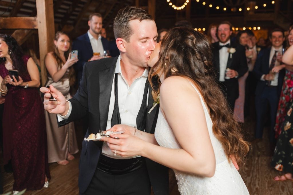 At a wedding reception at The Barns at Hamilton Station, the bride and groom, elegantly dressed, share a kiss while holding pieces of cake. Guests in the background are smiling and taking photos.