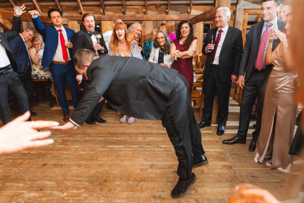 A man in a black suit bends forward on a wooden dance floor at The Barns at Hamilton Station, surrounded by people in formal attire, some clapping and cheering. The setting appears to be a joyous wedding reception.