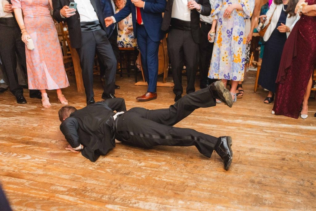 A person in formal attire lies face down on the wooden floor at The Barns at Hamilton Station while a group of people in varied clothing, perhaps guests from a wedding reception, stand around, observing.