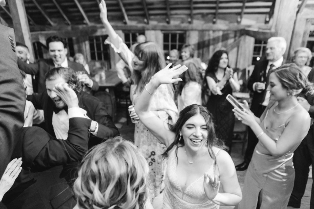 A group of people joyfully dance and raise their hands at a wedding reception. The focus is on a smiling woman in a dress at The Barns at Hamilton Station, while another person captures the moment on a phone.