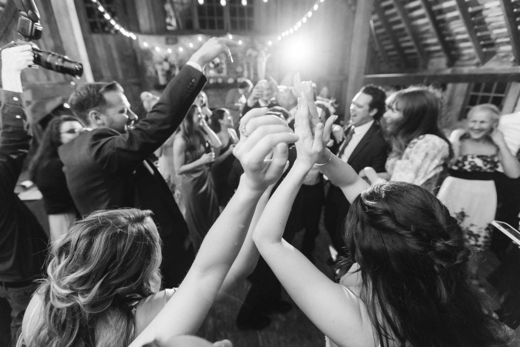 Black and white photo of people dancing joyfully at a wedding reception at The Barns at Hamilton Station, with hands raised and festive string lights overhead.