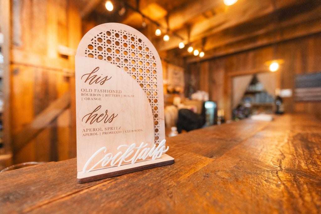 A wooden sign at The Barns at Hamilton Station details "his" and "hers" cocktail options, featuring an Old Fashioned and an Aperol Spritz, in a warmly lit rustic setting perfect for a wedding.