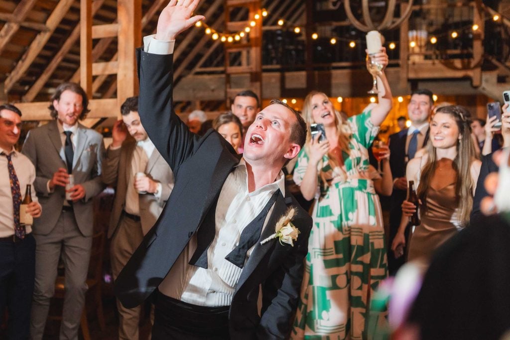 A man in formal attire enthusiastically raises an arm, surrounded by people in suits and dresses at a lively wedding reception. String lights decorate the wooden ceiling of The Barns at Hamilton Station.