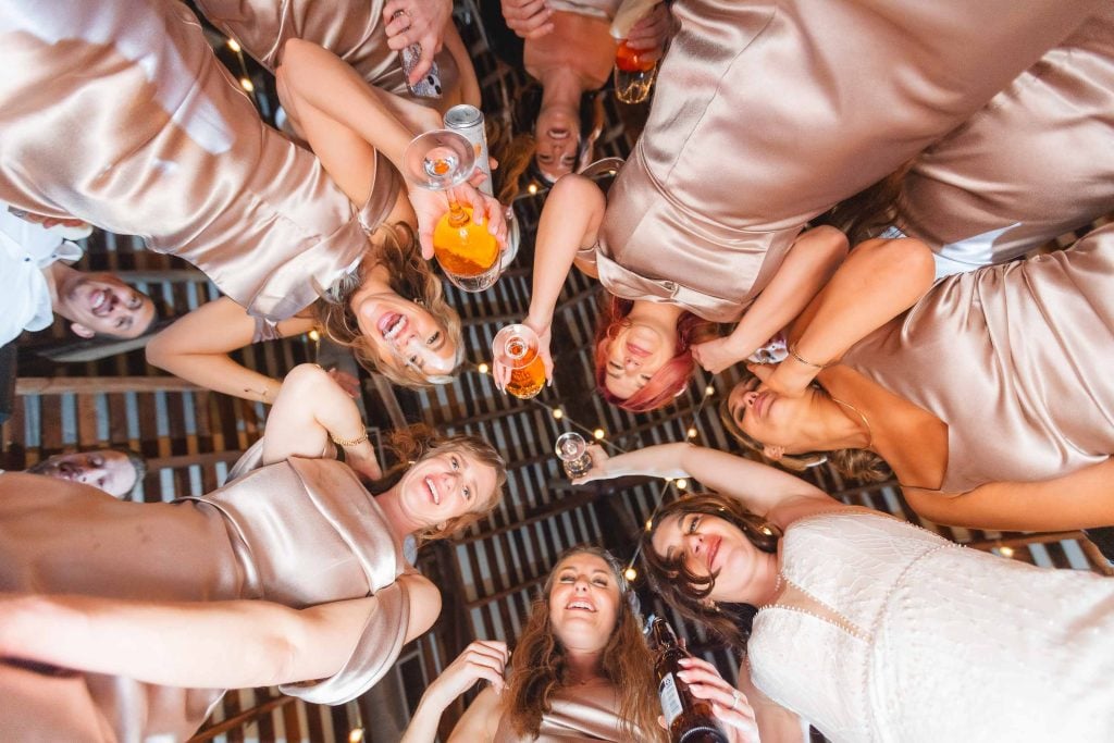 A group of women in champagnecolored dresses and one in white stand in a circle at The Barns at Hamilton Station, holding drinks and looking down at the camera with smiles and laughter during the wedding reception.