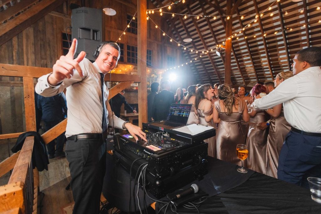 A DJ wearing a tie gestures to the camera while playing music at The Barns at Hamilton Station reception. A group of people in formal attire are dancing and socializing in the background under string lights, capturing the joyful essence of a wedding celebration.