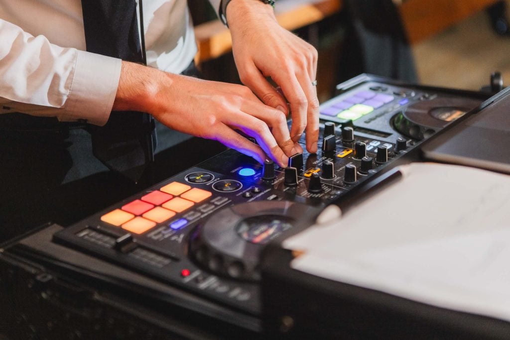 At a lively wedding reception at The Barns at Hamilton Station, a person adjusts controls on a DJ mixer, featuring brightly lit buttons and knobs.