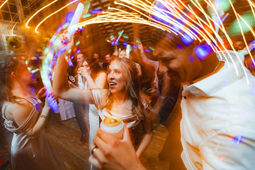 People are dancing at a lively wedding reception at The Barns at Hamilton Station, illuminated by colorful and swirling lights. One woman in the center is holding a drink and waving a light stick.