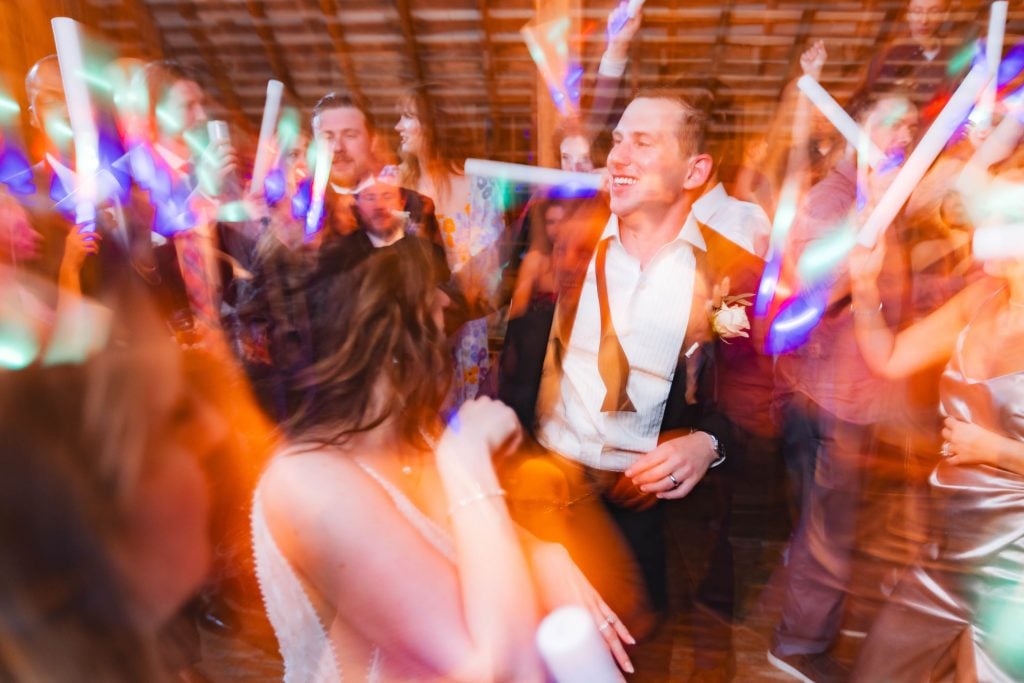 A group of people dance energetically at The Barns at Hamilton Station wedding reception, with colorful lights creating a dynamic and festive atmosphere.