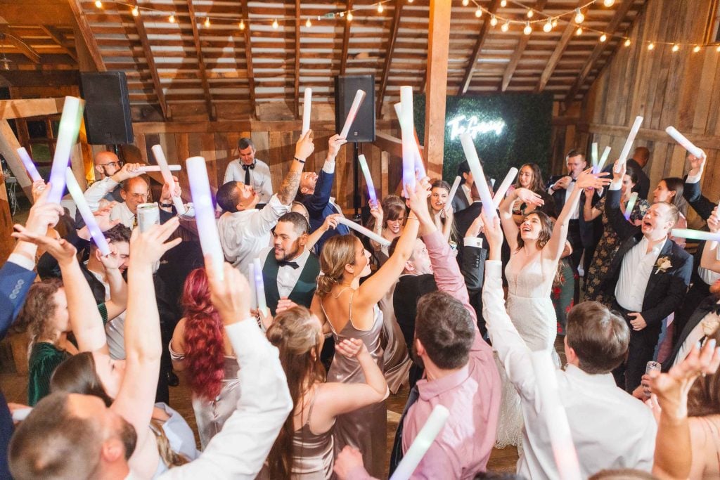 A lively wedding reception at The Barns at Hamilton Station with guests dancing, holding glowing sticks aloft, and having a joyful time under string lights.