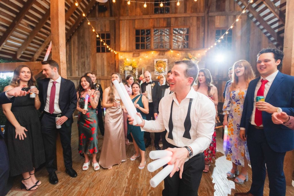 At The Barns at Hamilton Station, a man in suspenders energetically waves long white objects amidst a lively wedding reception crowd. Attendees in formal attire react with excitement and interest under the string lights hanging overhead.