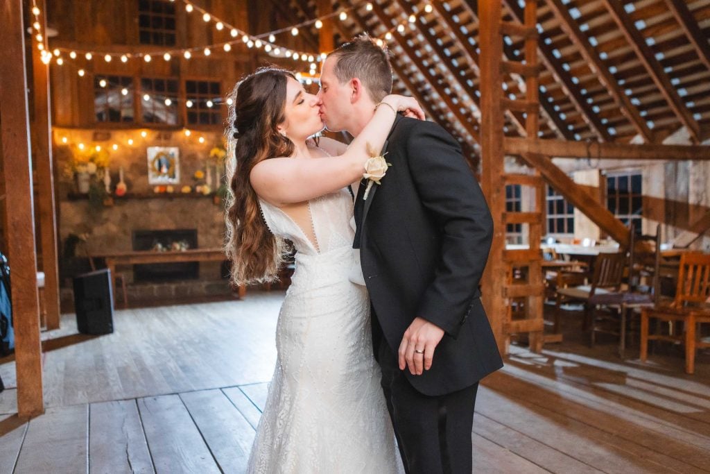 A bride and groom share a kiss on the dance floor of The Barns at Hamilton Station, a rustic venue adorned with string lights and wooden beams, perfect for an unforgettable wedding reception.