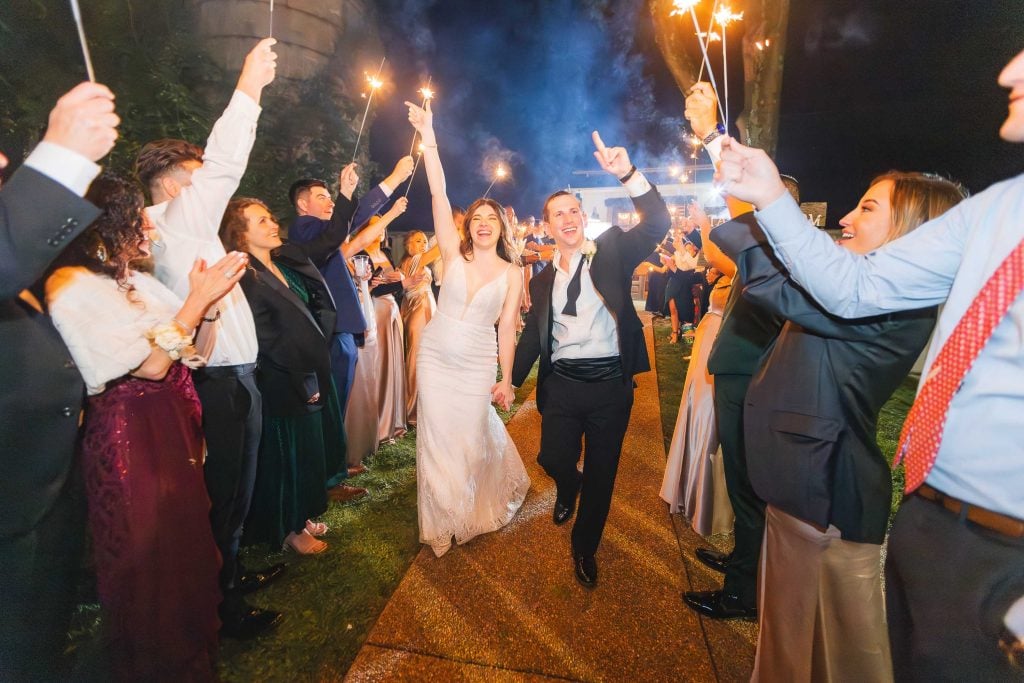 A bride and groom walk handinhand down a path at The Barns at Hamilton Station, smiling and celebrating, surrounded by guests holding sparklers at night.