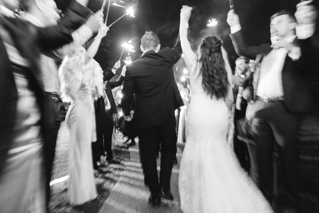 A blackandwhite photo of a bride and groom walking through a crowd holding sparklers, captured from behind at their wedding reception. Guests are celebrating, some with arms raised and sparklers lit, creating a magical atmosphere at The Barns at Hamilton Station.