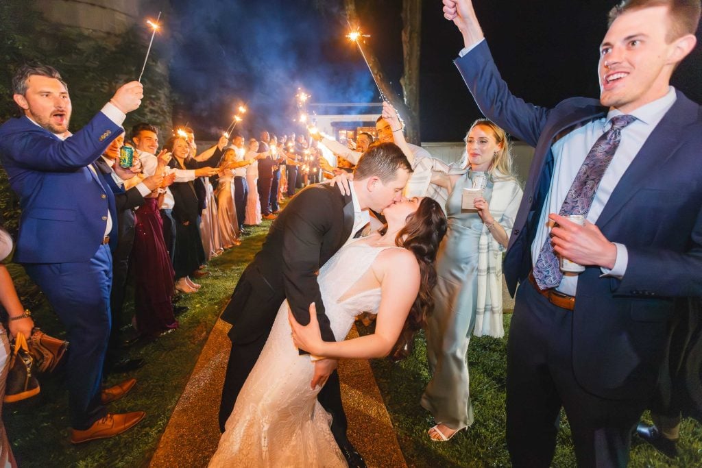 A bride and groom kiss at night, surrounded by guests holding sparklers at The Barns at Hamilton Station, celebrating their wedding reception in a festive atmosphere.