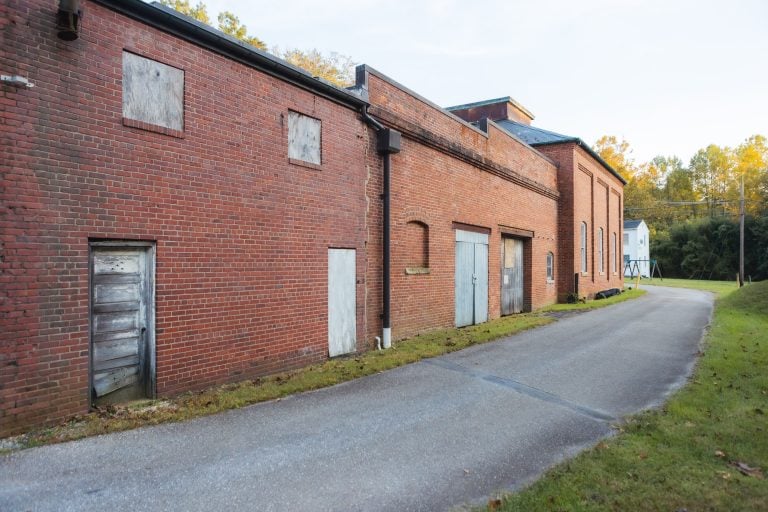 An old, boarded-up brick building stands quietly within Annapolis Waterworks Park, a paved road curving alongside it, surrounded by lush grass and towering trees.