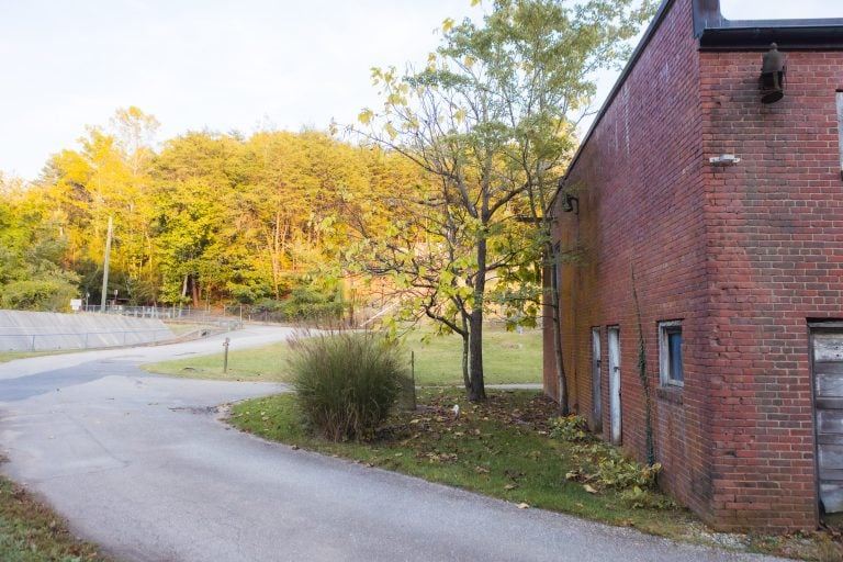 A narrow road winds past a red brick building in Annapolis Waterworks Park, bordered by grass and trees adorned with green and autumn-hued leaves.