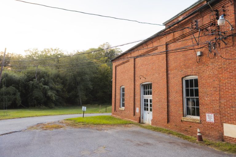 An old brick building with large windows and a wooden door, surrounded by utility poles and wires, sits next to a quiet road near Annapolis Waterworks Park, framed by lush green foliage.