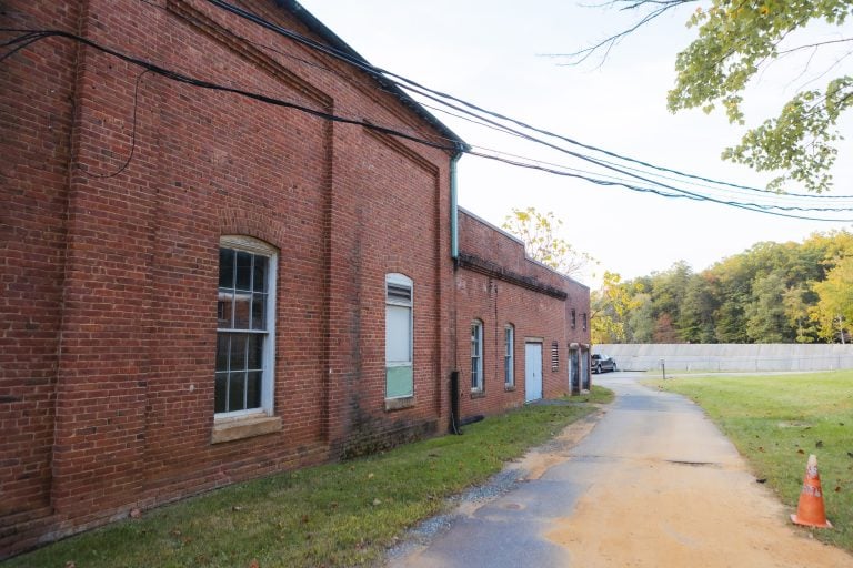 A brick building with several windows stands beside a narrow paved path in Annapolis Waterworks Park. Power lines run above, and a traffic cone is seen on the path. Trees and a fence are visible in the background, adding to the park's serene setting.