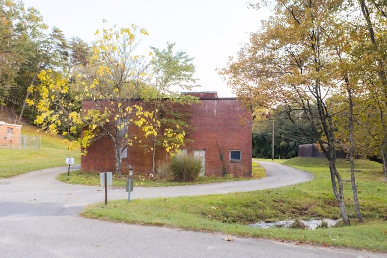 Nestled next to a winding road, a small brick building with minimal windows is enveloped by trees with yellowing leaves, forming an enchanting entrance to Annapolis Waterworks Park.