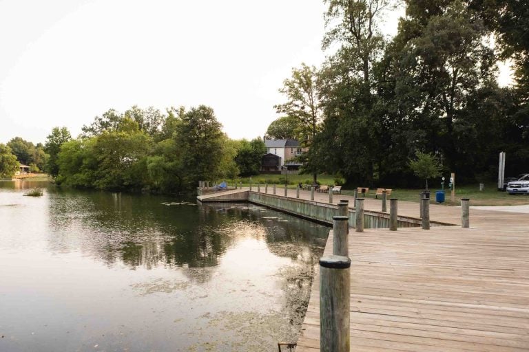 A wooden dock extends over the calm waters of Lake Elkhorn, surrounded by trees and a small building. Benches line the dock under a cloudy sky.