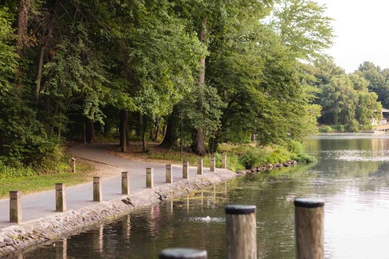 A paved path runs alongside Lake Elkhorn, a tree-lined lake with several wooden posts along the water's edge.