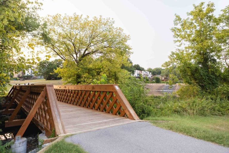 A charming wooden footbridge spans a small creek, embraced by green trees and grass on either side, guiding the way toward a distant neighborhood near Lake Elkhorn.