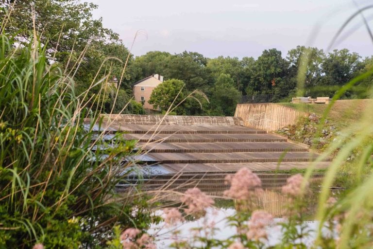 A stepped dam with water flowing down is surrounded by lush greenery, part of the picturesque Lake Elkhorn. A house is visible in the background, nestled among trees.